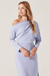 A woman is wearing a light blue off-the-shoulder dress with long sleeves. The dress has a relaxed, draped fit around the shoulders and a fitted waist. The cuffs of the sleeves are slightly darker in shade. She has long, wavy, light brown hair.